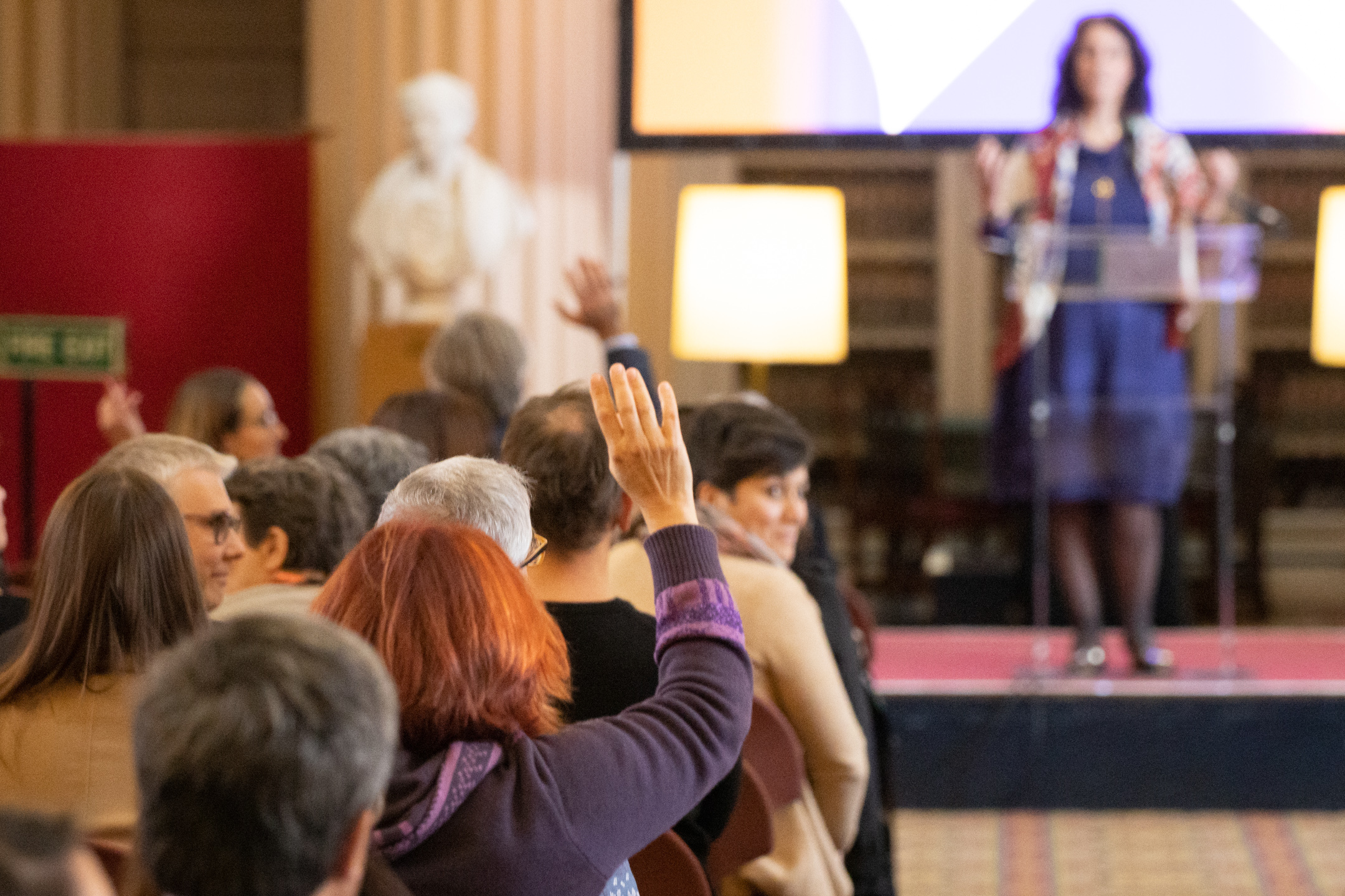 Woman stood on a stage with an audience sat in front. One member of the audience is looking back towards the camera, and another has their hand up.