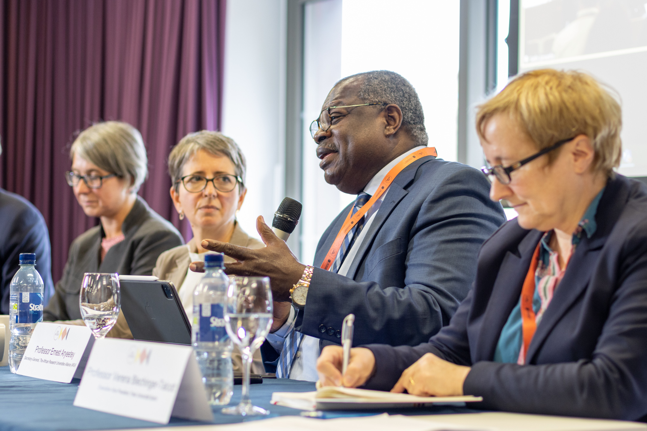 Four people sat at a table with name signs in front of them. One of the speakers is holding a microphone and talking.