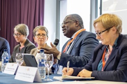 Four people sat at a table with name signs in front of them. One of the speakers is holding a microphone and talking.