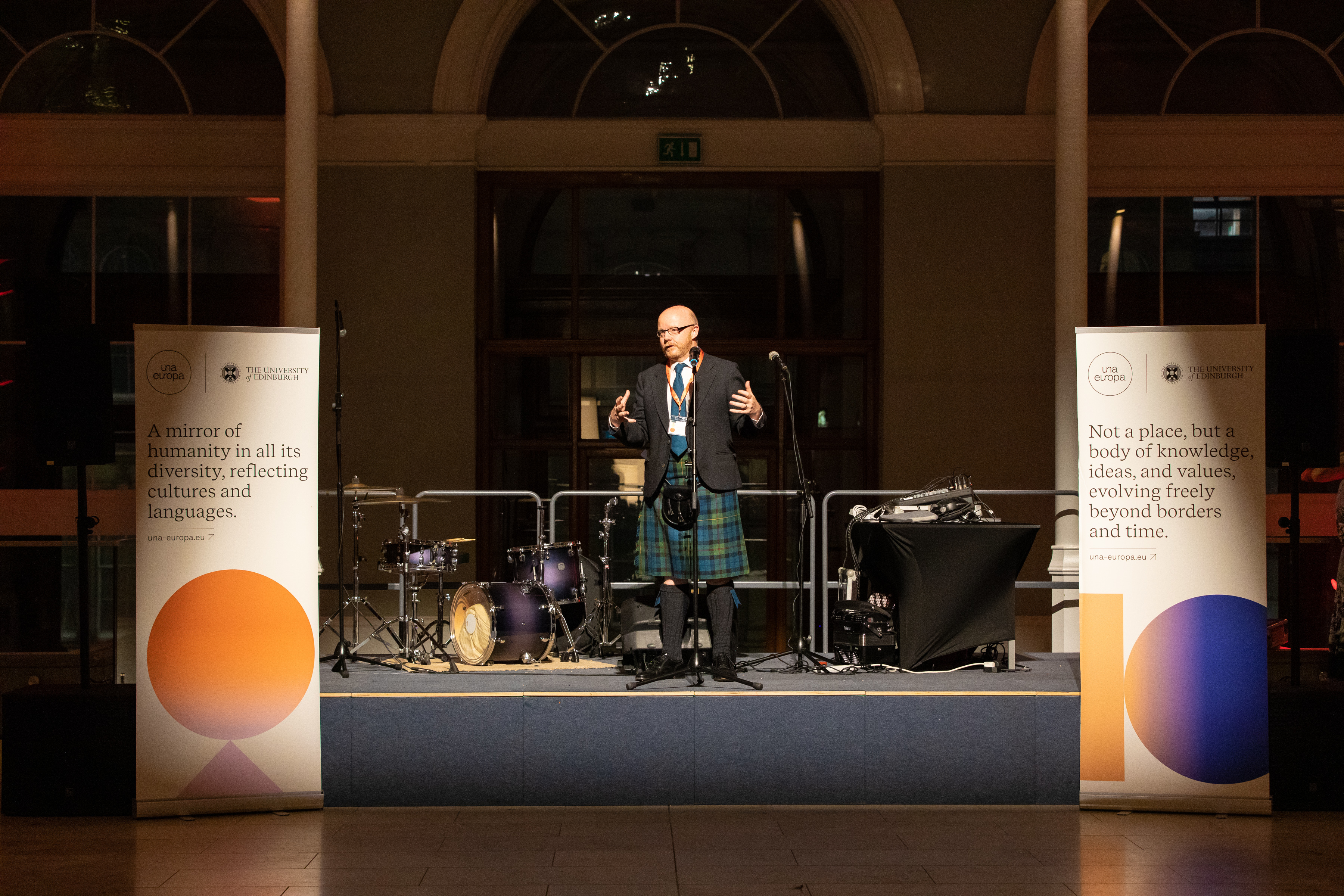 Man wearing a kilt and traditional Scottish dress with arms outstretched, flanked by two pull-up banners with a colourful shape design.