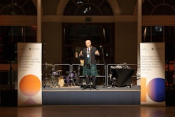 Man wearing a kilt and traditional Scottish dress with arms outstretched, flanked by two pull-up banners with a colourful shape design.