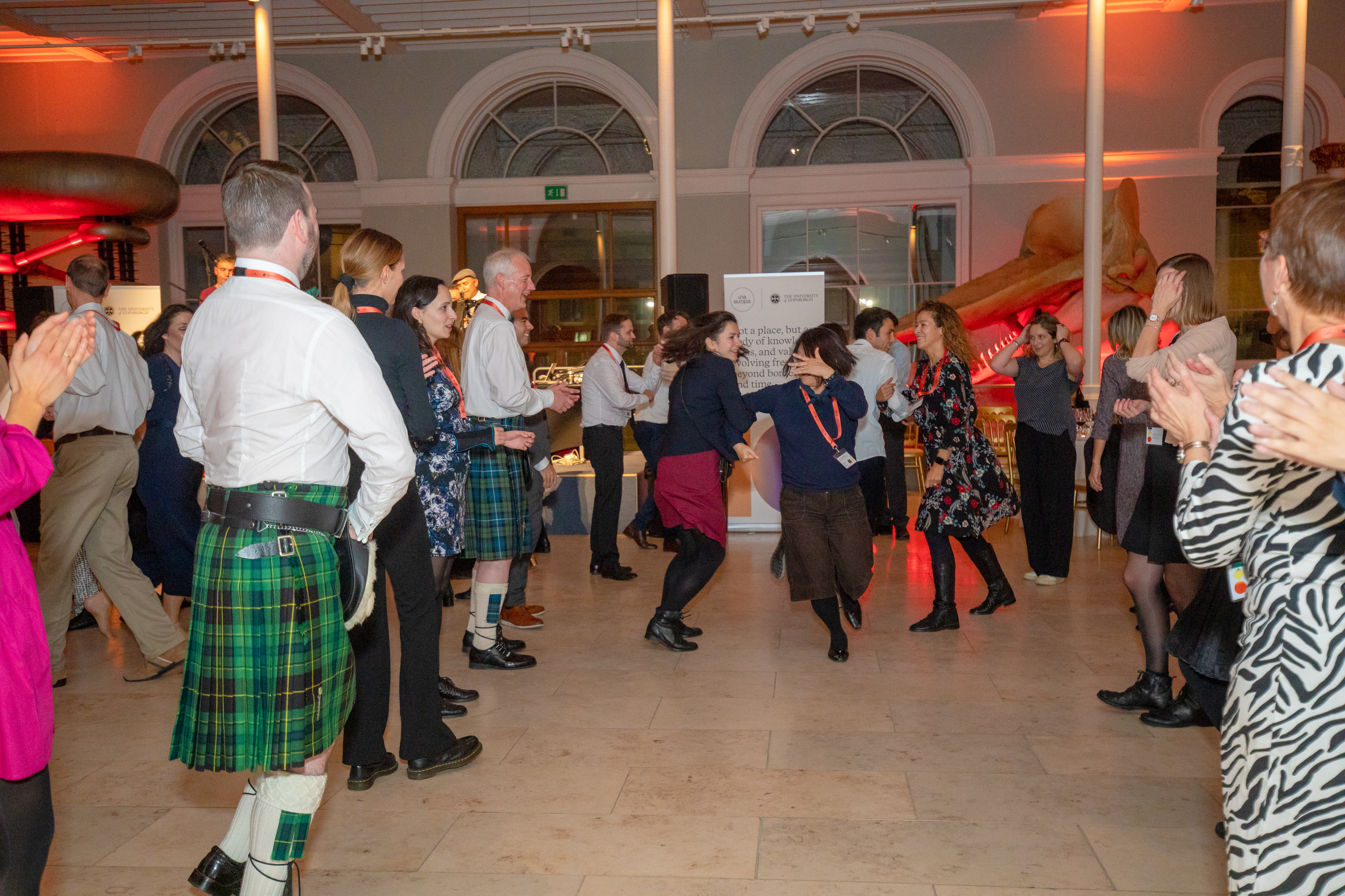 People dancing a traditional Scottish ceilidh, with arms hooked in each other and wearing traditional Scottish dress including a kilt