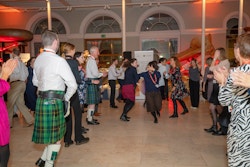 People dancing a traditional Scottish ceilidh, with arms hooked in each other and wearing traditional Scottish dress including a kilt
